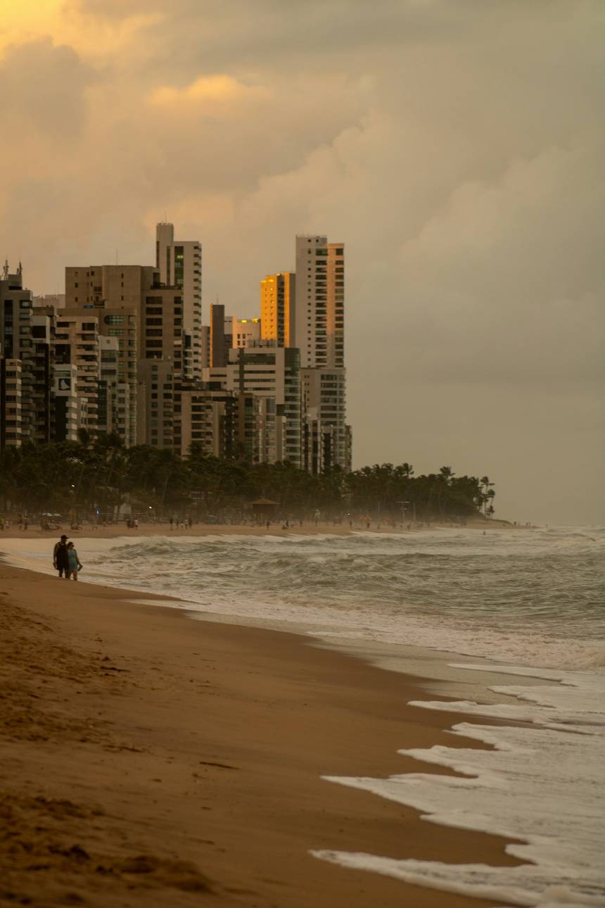 Orla com prédios altos e faixa de areia, com o mar à frente, ao entardecer.